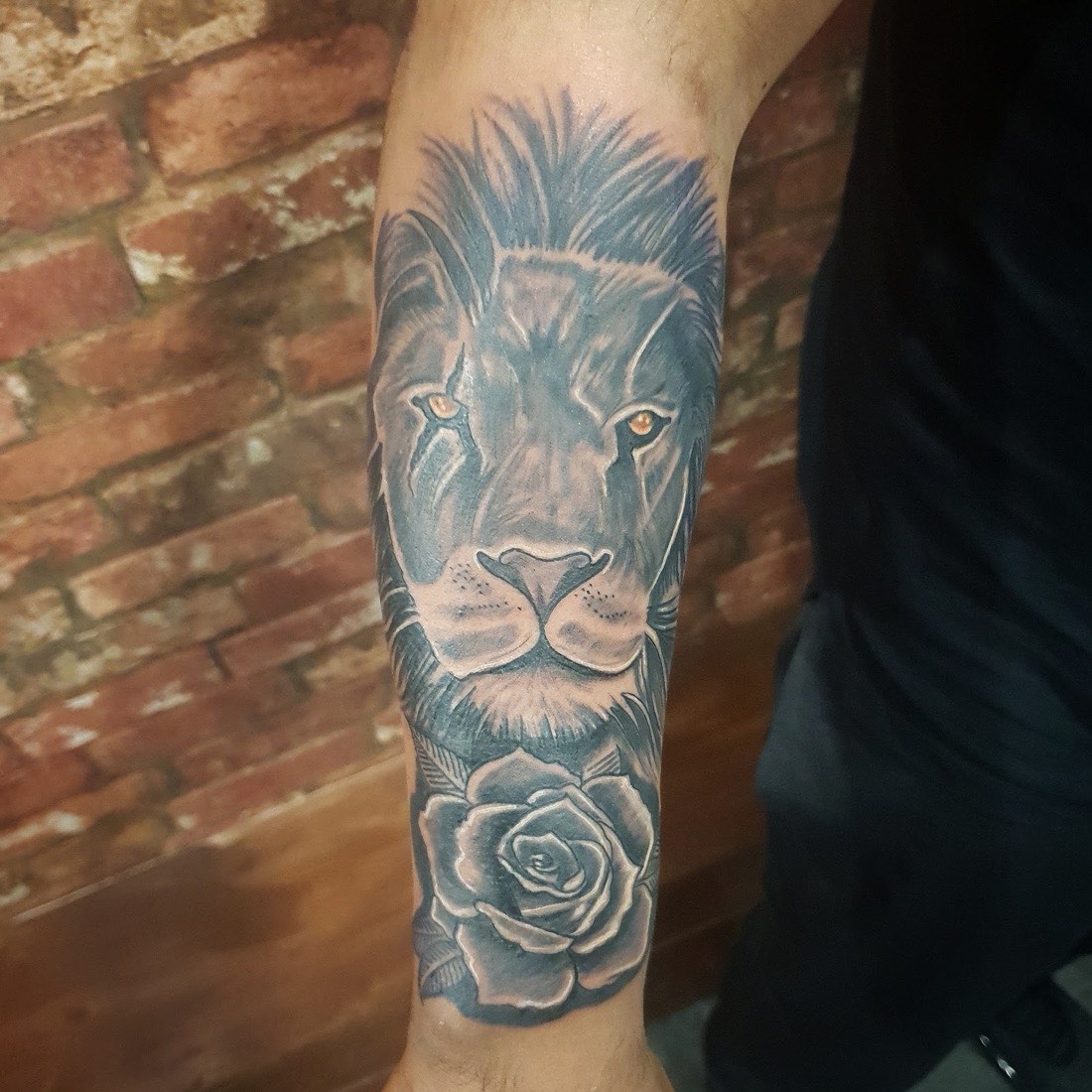 Realistic lion and rose tattoo in black and grey against a brick wall, by Scorpion Ink